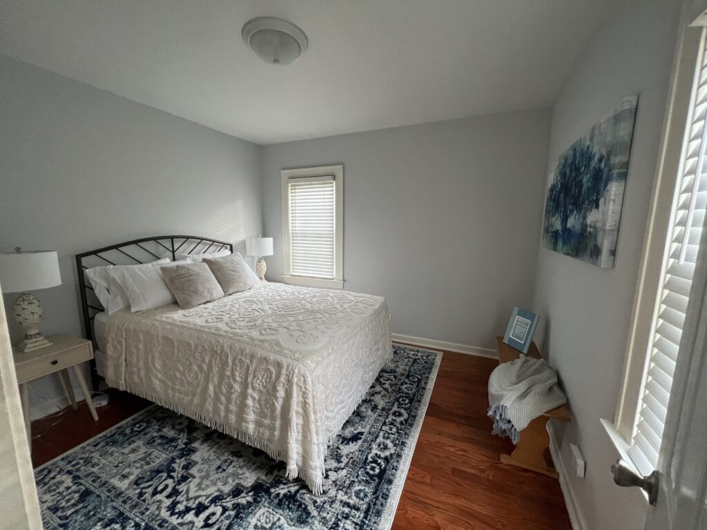 Beach Town Getaway image of the queen bedroom. Tan and white bedding, lamps, and bedside tables, withe blue accents in the rug, painting, and other decor.